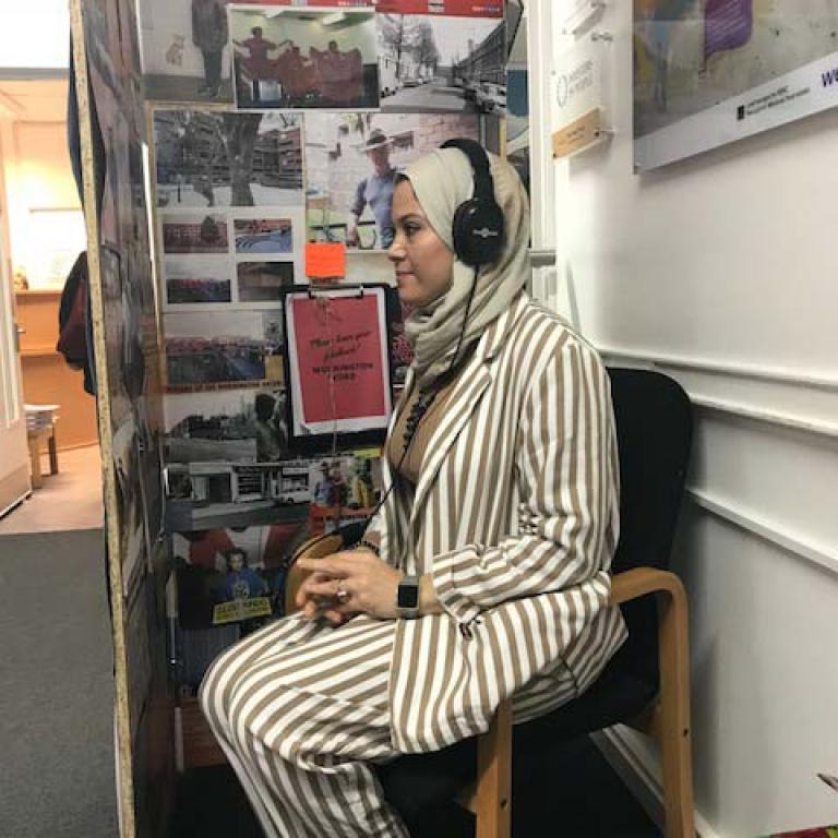 Inside The Westway Trust main reception at Thorpe Close, W10, a woman sits inside the Oral History Booth. She listens to the recorded stories of Wornington Green Estate residents whilst seeing photographs taken across the estate. Photo taken by Natasha Langridge 2019.