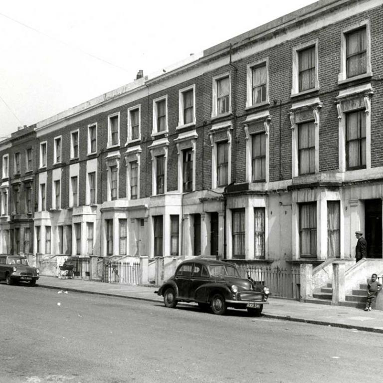 A black and white photo of Wornington Road, looking east before Wornington Green Estate was built, depicting Victorian terrace houses. There is a young boy leaning against a wall as a man comes down the front steps behind him. A pram stands further up the pavement. There are three cars parked on the empty road. Photo courtesy of RBKC Local Studies and Archives, 1970.