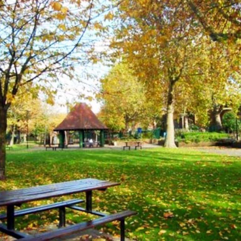 The original Athlone Gardens in Autumn. A brown table with attached benches sits on the grass to the fore. A circular pavilion with a brown roof, known as the ‘witches hat’ to locals, stands in the background. Tall trees with abundant yellow and orange leaves edge the central expanse of green lawn. Photo reproduced from The United Estate of Wornington Green, 2008.