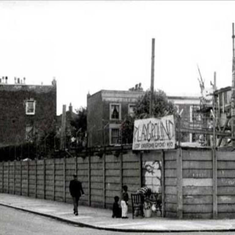 Photo depicts the Notting Hill Adventure Playground from Wornington Road on the south side. On this corner side the playground has a wooden fence with a big sign saying playground attached to the chain link fence atop. In front, on the pavement, is a man painting a mural onto the fence with two young children watching on. Behind in the playground you can see a big higgledy piggledy wooden structure with a boy climbing up one side. To the left on the street and further behind are residential houses.