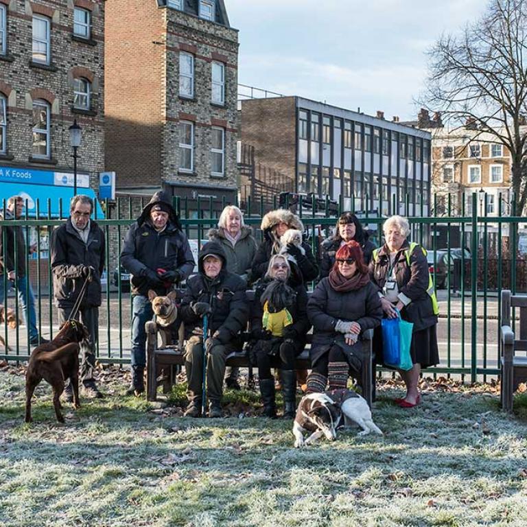 A frost covered grass to the fore. In the centre are nine members of the famous dog walking club sitting on and standing around a bench in coats and scarfs. Three dogs are close by. One called Fidel shares the bench with them as they pose for the camera. A green railing fence is behind them above which is seen AK Foods on Portobello Rd and Lloyd Williamson School on Telford Rd. Photo by Kevin Percival 2019.