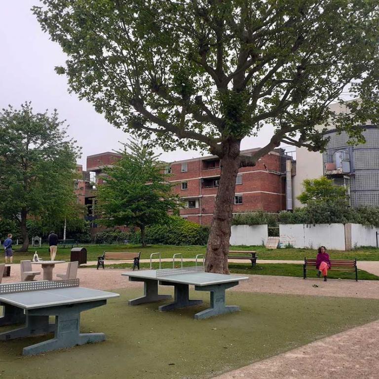 A view in Athlone Gardens from the Portobello Road side. Newly laid table tennis pitches stand beneath a leafy mature tree. Two other trees stand in the background in front of the red bricked Chesterton and Chiltern Houses. To the right is the fenced off and demolished Lionel House walkway and the side of the glass entranceway to Watts House. In the centre a woman sits on a park bench placed on a neat pathway. Photo by Constantine Gras 2020.