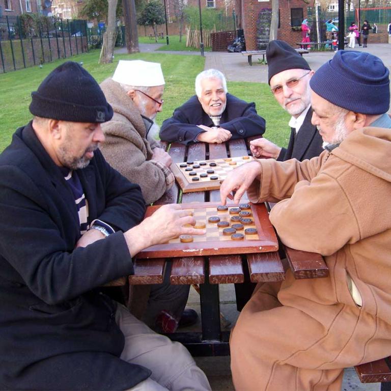 To the fore five men play dominoes as they sit around a brown table. Two look towards the camera. Three are deep in concentration. To the right is the lower half of Wheatsone House. A concrete path winds though the centre. A blackboard leans against a brick wall to the left in the background. Photo reproduced from The United Estate of Wornington Green, 2008.