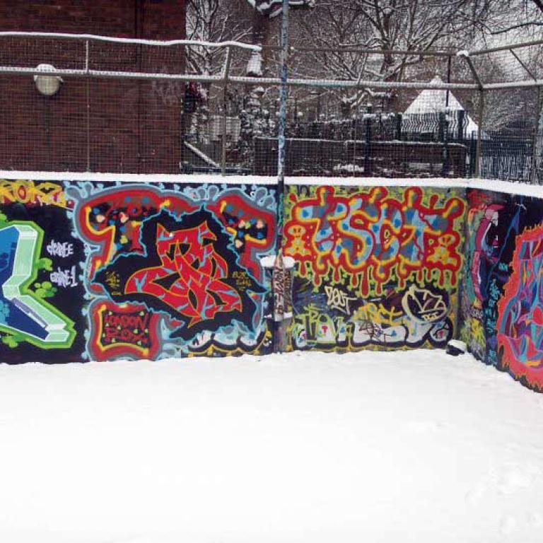 Snow covers the floor of the basketball court highlighting the multi-colours of the two graffitied walls. A metal fence rises above the back wall. Trees and the snow covered cone shaped roof of the witches hat in the original Athlone Gardens can be seen through it. Photo by Junior Tomlin 2012.