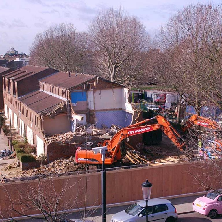 Picture of a partially demolished Faraday House revealing the inside blue wallpaper of a residents former home.  In the foreground rubble, is an orange demolition machine. In the background are trees from the original Athlone Gardens. Photo taken from window of glass and jewellery workshops in Wornington College. Photo by Ken MacDonald, 2011.