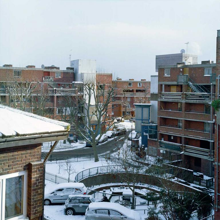 Looking South-East across the estate from Thompson House towards Katherine House. From l to r: Pepler House (left foreground), Katherine House (middle background), Wells House and the community garden, and Rendle House. The ground is covered in snow, on a bright winters day, and a resident clears her car windscreen in the shadow of the buildings in the foreground. Photo by Kevin Percival, a resident of Pepler House, 2018.