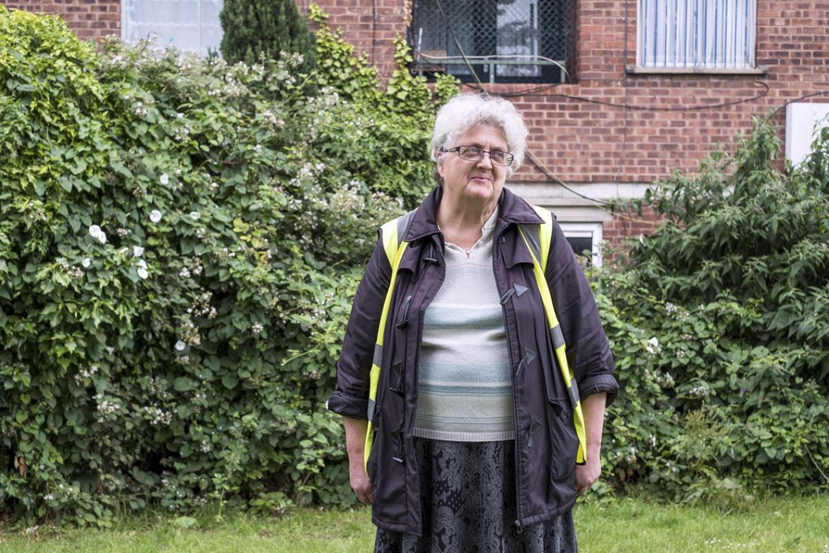 Kathleen is standing ouside with big thick bushes and a red brick building behind her. Kathy has white hair and black framed glasses and is wearing a dark purple jacket with toggles with a high res sleevless jacket over the top. Underneath she is wearing a fine knit top with light stripes of green and blue and a dark coloured skirt with a light printed pattern on top.
