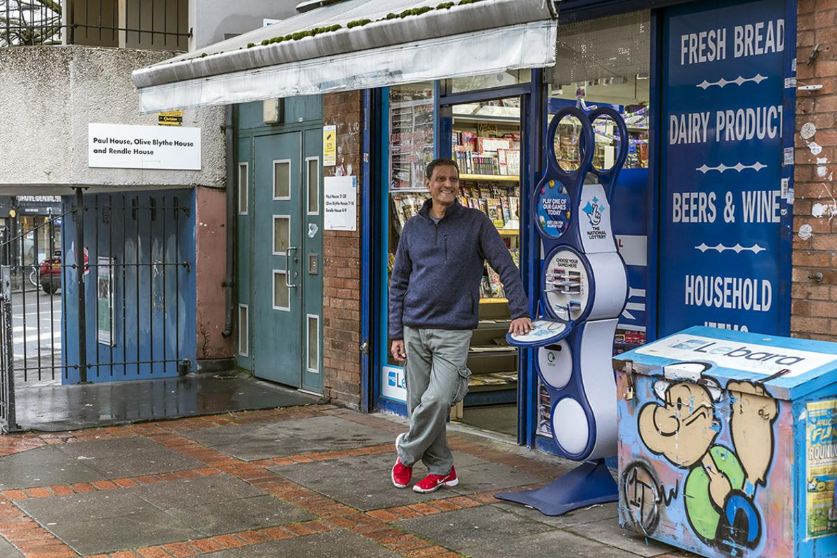 Kirit is standing outside his newsagent shop. There is a taupe coloured door with panels in it to his right and black railings, one end attached to a blue wall. Kirit is leaning on a lotto stand-alone unit with a large padlocked metal box next to it painted with a picture of Popeye. Kirit is wearing a blue collared sweatshirt, light grey commando trousers and red trainers. He has short dark hair.