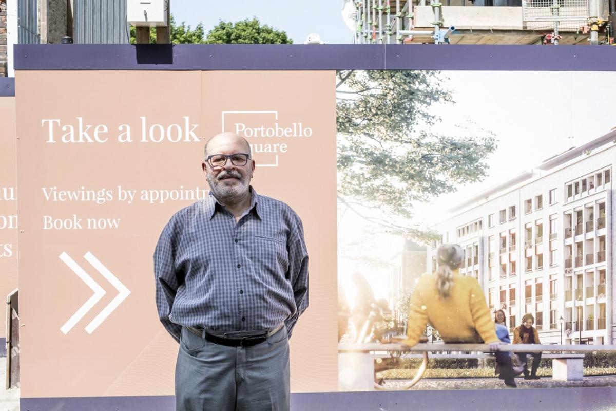 Moshen is standing in front of a huge billboard depicting the new buildings of Portobello Square. He is bald with a grey beard, a grey moustache and black framed glasses. He is wearing a purple checked shirt and a pair of grey suit trousers with a black belt.