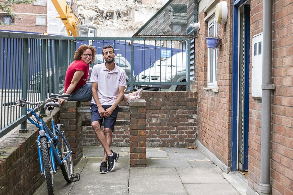 Latifa and Rachid are outside their ground level flat in the courtyard. The blue railings are on street level, the floor is concrete and there is a red brick barbecue unit. A bicycle is chained to the railings. Latifa is wearing a red t-shirt, casual blue trousers, flip flops and she wears glasses. Rachis is wearing blue shorts, a polo t-shirt and black trainers.