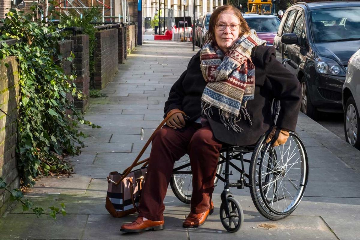 Margaret is in a wheelchair on a pavement with parked cars to her right. She is wearing a striped wool scarf, a black winter jacket, brown leather gloves, brown trousers, brown shoes, a pair of glasses with a handbag looped over one hand.