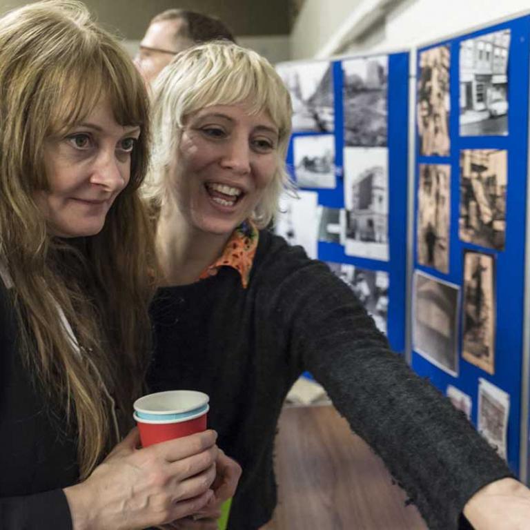 Photo depicts two woman looking and smiling at a photo in a montage of photos mounted on a blue board