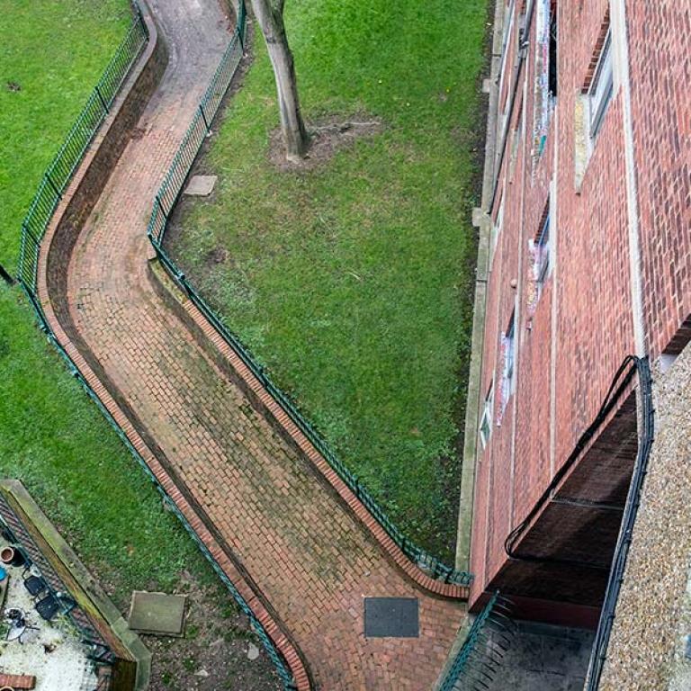 A view from the 4th floor walkway of the garden square below that is situated between Katherine House, Macaulay House and Breakwell Court. A red brick path leads through the grass. To the left is a residents garden with several brightly coloured flower pots. Photo by Kevin Percival, a resident of Peplar House.