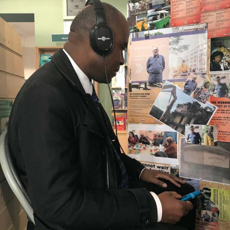 Inside North Kensington Library, a suited man, listens intently to oral history extracts from the Wornington Green Estate. The booth also has photographs of people and places across the Wornington Green Estate. Photo taken by Natasha Langridge 2019.