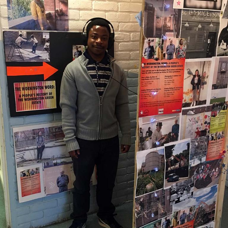 James Oluwaseye Ogundipe, staff member at the Venture Community Centre, listening to the Wornington Word Oral History Booth. The booth allows people to interact with the recorded stories of residents whilst seeing photographs taken across the estate. Photo taken by Micky Pallant 2019.