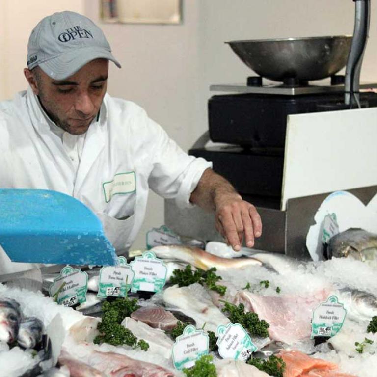 A photo of a fishmonger in Golborne Fisheries on the Golborne Road. He wears a white apron and is scooping ice over haddock, salmon, halibut, tuna and plaice on the counter. A metal scale is in the background. Photo taken by Darnell Da Costa, 2019.