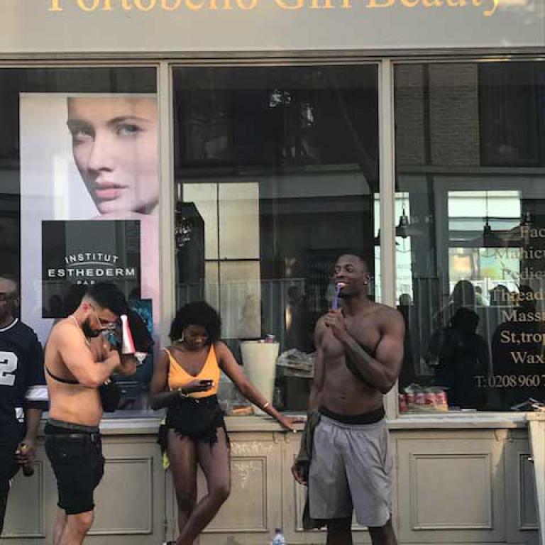 Photo depicting a shop at 355 Portobello Road, Portobello Girl Beauty, a beauty salon. Leaning against its glass front, a girl in shorts and an orange t-shirt checks her mobile phone while two men on either side of her, also in shorts, blow woofer horns during carnival. Photo by Natasha Langridge, 2019.