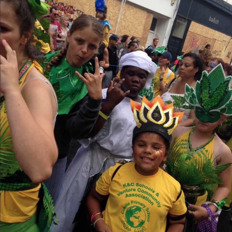 Photo depicts group of five children from the Venture Centre who are taking part in the Notting Hill Carnival on Portobello Road. Several are in green and yellow costume. One girl is wearing a white dress. The smiling girl in the foreground has a flamed head dress with a yellow t-shirt advertising Kensington and Chelsea Schools and Venture Community Association. Photo reproduced from @VentureCentre1 Twitter feed, 2016.