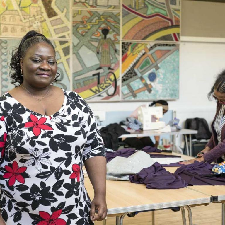 Photo of Esther Mangoung who is the teacher of the Sewing Club at the Venture Centre. She is standing in the foreground wearing a colourful black and red floral dress. In the background are four women working at sewing machines and cutting fabric. On the wall is a large African mosaic art work. Photo by Kevin Percival, 2018.