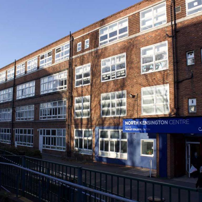 Photo depicts the exterior facade and blue signage of North Kensington Centre, part of Morley College. This was formerly Kensington and Chelsea College and Issac Newton Upper School. Dappled light falls across the red brick building and its large latticed windows. Photo by Hannah Hutch, 2020.