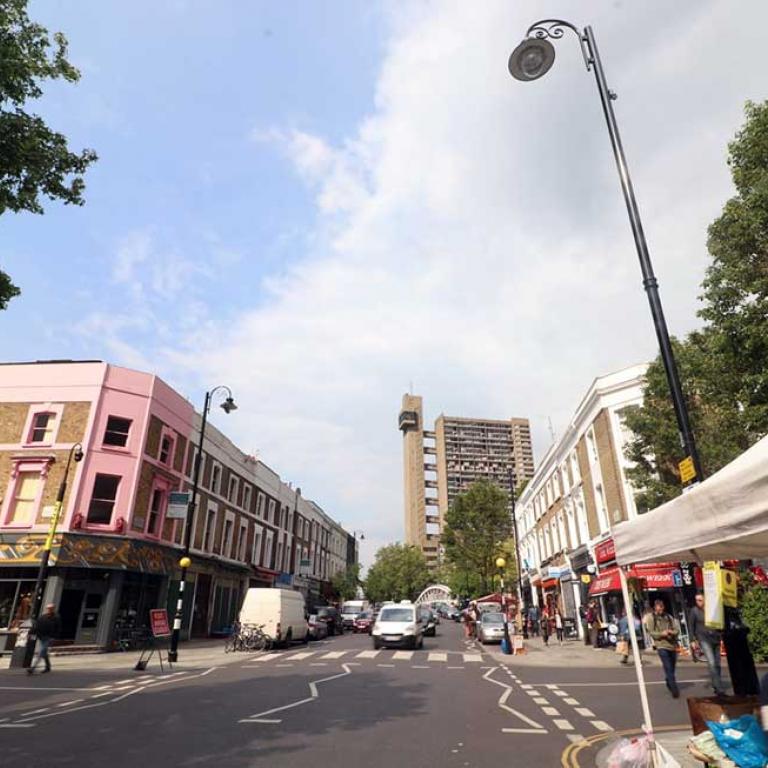 Photo depicts an extreme wide angle shot of Golborne Road, looking north towards Trellick Tower. The white canopy of a flower stall is in the foreground right. Cars in the distance approach a pedestrian crossing between the rows of shops. There are Road Ahead Closed signs to the left at the junction of Wornington Road and Golborne Road. Photo taken by Darnell Da Costa, 2019.