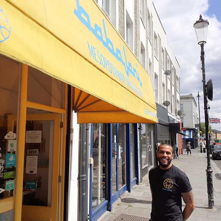Photo depicts a smiling Kris Parker on the pavement of Portobello Road outside Babajani, his Mesopotamian restaurant/cafe. A yellow awning sits above the shops glass frontage. In the background Union Jack bunting hangs across the street between buildings. Photo by Constantine Gras, 2020.