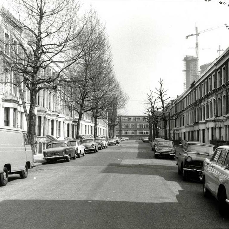 A black and white photo of Wheatstone Road, looking east before Wornington Green Estate was built, depicting Victorian terrace houses. The road has tall trees, several pollarded, with numerous cars parked in the street. At the end of the road is Issac Newton Upper School on Wornington Road and in the background is Trellick Tower under construction with several cranes. Photo courtesy of RBKC Local Studies and Archives, 1970.