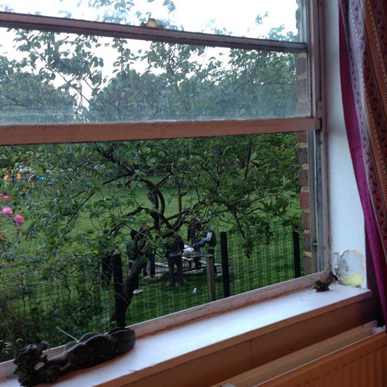 View of Athlone Gardens from a window at Wheatstone House. An ornamental dragon sits inside on the white window ledge. Through the half open window, there is a late blooming cherry tree and a group of men seen standing in discussion on the expanse of green grass beneath its curly branches. Photo by Natasha Langridge, 2014.