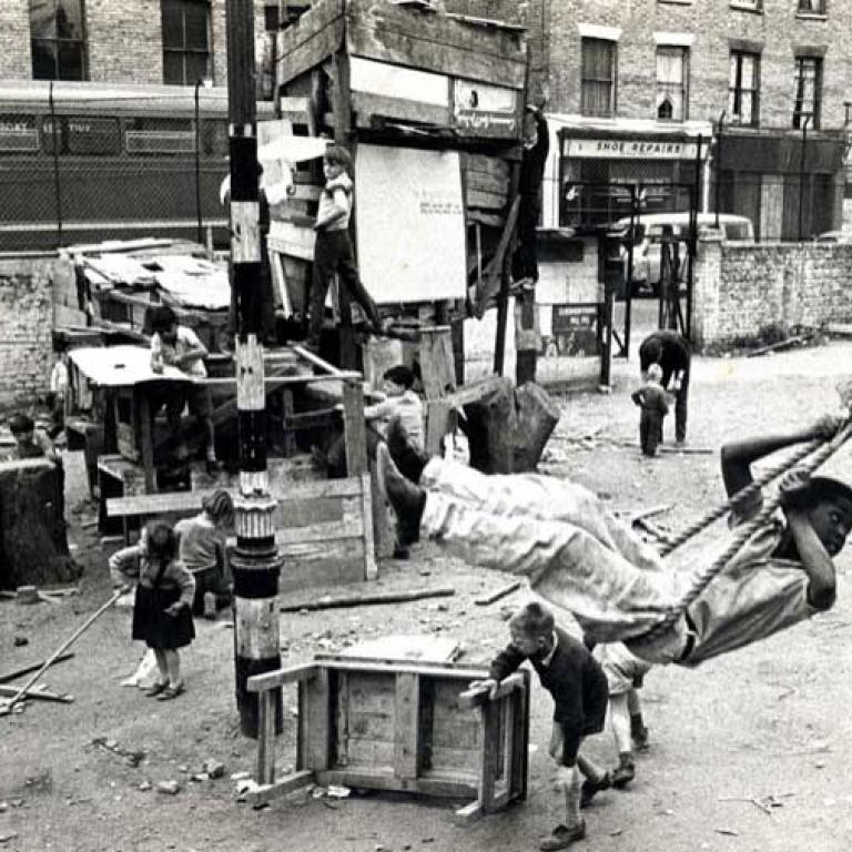 Photo depicts the Notting Hill Adventure Playground circa 1950s. Children playing, a young boy in the foreground is swinging on a rope swing, in the background is a higgledy piggledy wooden structure with two boys climbing on it. Behind is a chain link fence and then behind that a row of residential houses with shops on the ground level, one is a shoe repairer and a passing bus.
