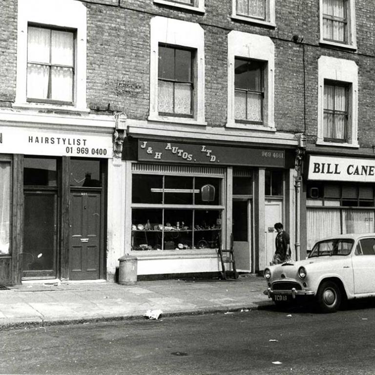 A black and white photo of Wornington Road depicting three shops: Henson Hairstylist, J&H Autos Ltd and Bill Cane Turf Accountant. A light coloured car is parked outside with a woman walking past carrying shopping bags. Photo courtesy of RBKC Local Studies and Archives, 1970.