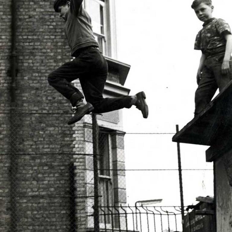 Photo depicts a corner of the Notting Hill Adventure Playground circa 1950s. In the foreground a young boy is mid flight after jumping off the roof of a hut whilst holding an open umbrella above his head. Another boy watches on from the roof and another boy is standing on the ground next to the hut. In the background is a chain link fence behind which is the side of a residential house and a streetlight in front.