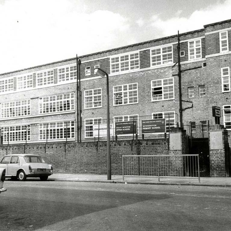 A black and white photo of Issac Newton Upper School and Wornington Primary School on Wornington Road. The building is an imposing structure of four floors with large latticed windows. The school gate is closed and to the right three boys walk behind a parked car. Photo courtesy of RBKC Local Studies and Archives, 1969.