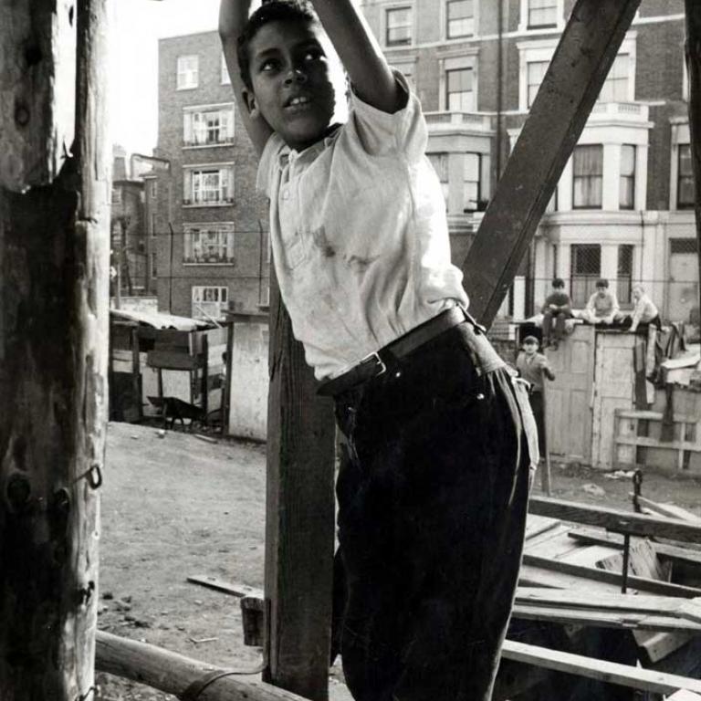 Photo depicts the Notting Hill Adventure Playground circa 1950s. In the fore-ground there is a young boy standing on a higgledy piggledy wooden structure, his arms are stretched up holding onto a wooden beam above. In the background there are three children atop another wooden structure, a bit like an old wardrobe, with a forth child on a pair of stilts in front of it. Behind is a chain link fence and a row of residential houses.