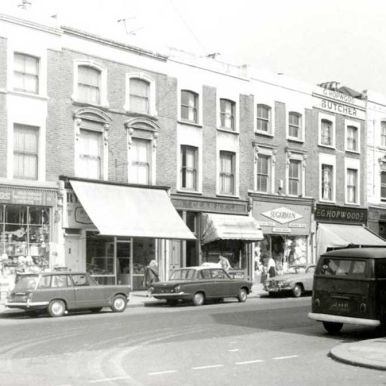 A black and white photo of Golborne Road's north side with the following shops: O’Mahony Bros (domestic store), Clarke (corn chandler), J Sugarman (ladies outfitter) and E. G. Hopwood (butcher). Several cars are parked on the street including a camper van. Photo courtesy of RBKC Local Studies and Archives, 1969.