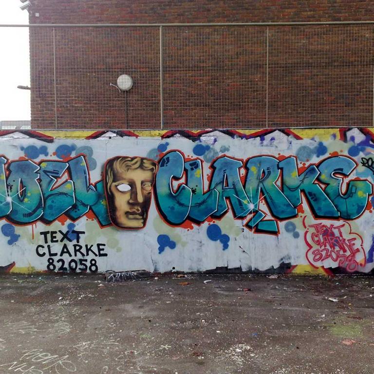 'Vote Noel Clarke' is graffitied in blue letters on a white wall in the basketball court that once stood behind Althone Gardens. Between Noels first and second name is a gold graffitied BAFTA award. Beneath that in black letters reads text 82058. To the front is the grey concrete court floor. Above is a silver fence. Photo by Junior Tomlin 2012.