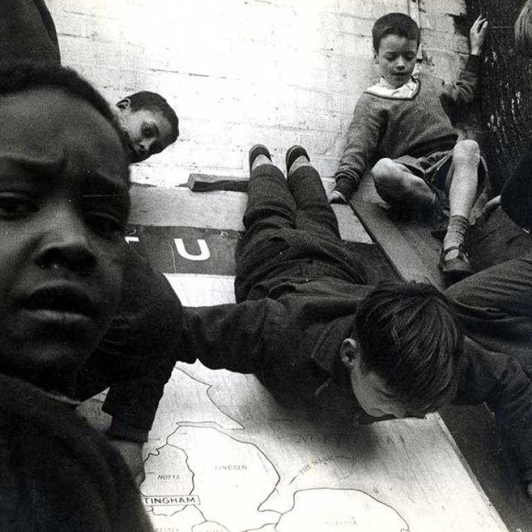 Photo depicts a group of five boys playing with a huge map of England that is leaning against a wall. One boy is sliding down the map while two sit at the top, to the right, watching him. Two other boys have turned to face the camera. Photo courtesy of RBKC Local Studies and Archive, circa 1960s.