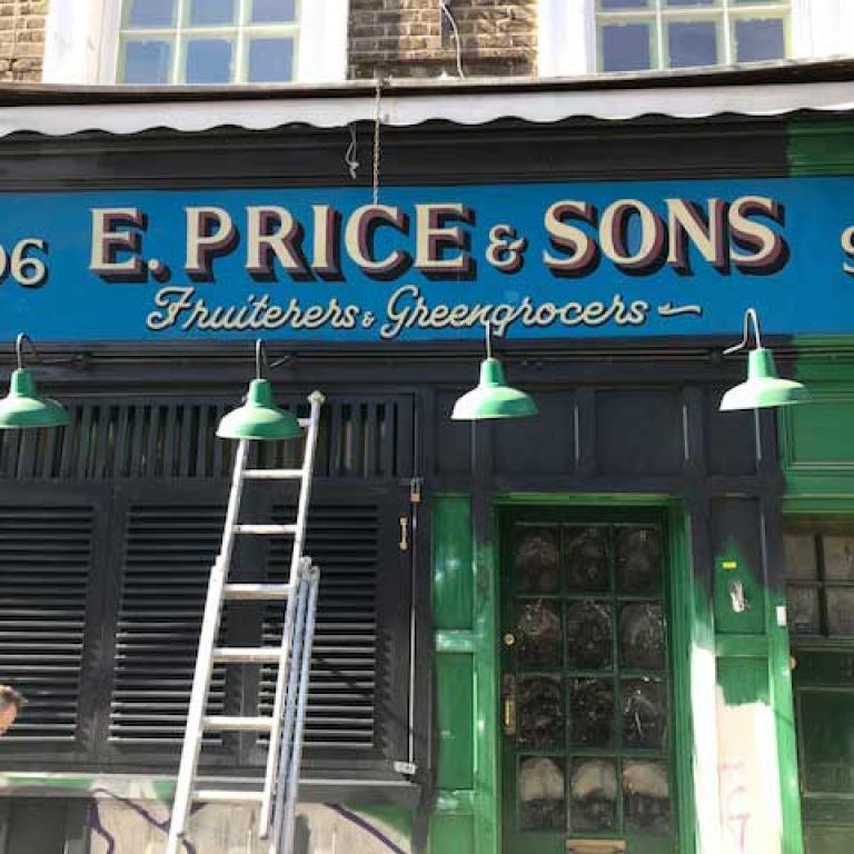 Photo depicting a shop at 96 Golborne Road. At the top of the photo is the old fashioned signage saying E. Price & Sons, Fruiterers and Greengrocers in gold lettering on a blue background. Four green lamps hang over the closed shop front and black shutters. A silver ladder leans against the shutters. E Price & Sons was run as a family business since 1937, but has now been sold. Photo taken by Natasha Langridge, 2020.