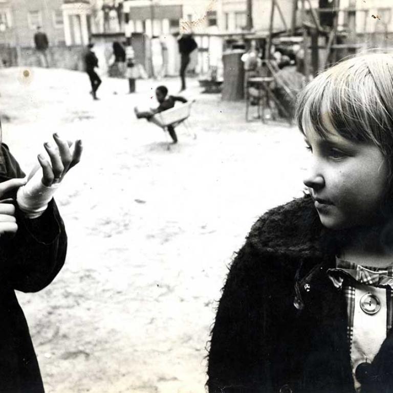 Photo depicts two girls in the Venture playground, one is looking at her bandaged hand and the other is lost in thought. In the background a small boy dances with a wheelbarrow in front of a group of teenagers. Photo courtesy of RBKC Local Studies and Archive, circa 1960s.