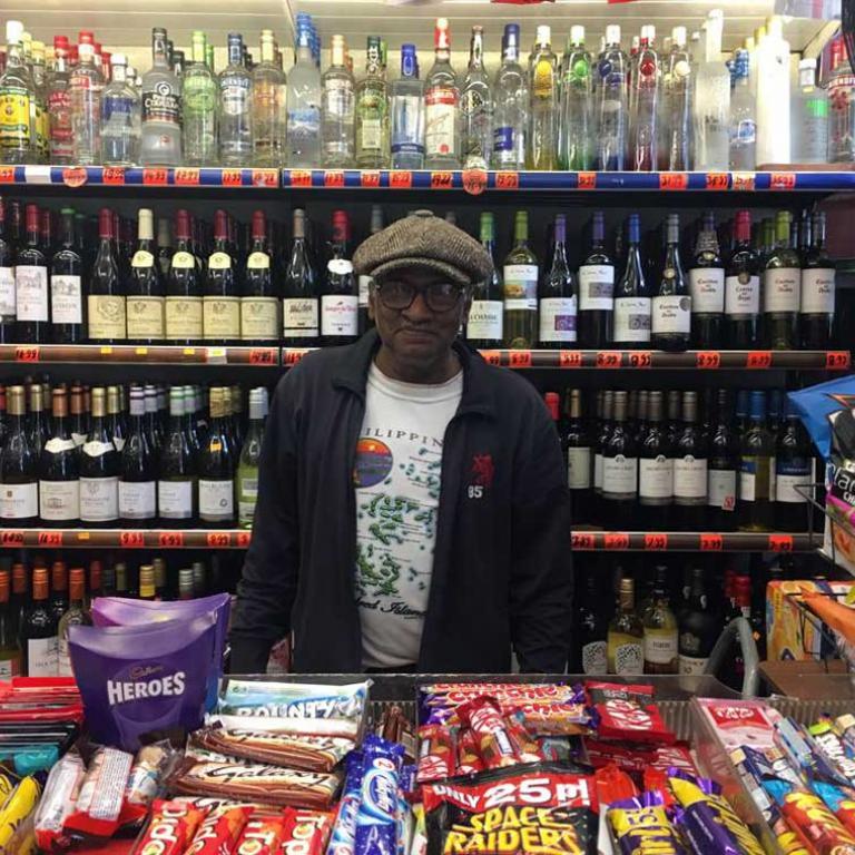 A photo of the owner of Cut Price Liquor Store on Golborne Road wearing a brown cap. He stands behind a counter of confectionary and in front of a display case of wine and spirits. Photo by Tedra Clover, 2019.