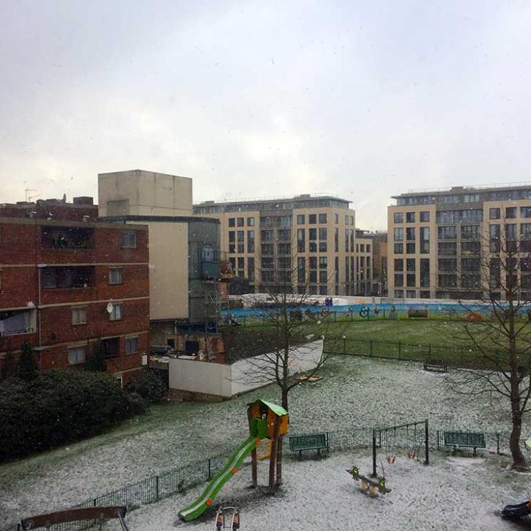 View of Althone Gardens from Chesterton House on a snowy day. Tiny snowflakes fall in front of the camera lens. To the left are the windows and balconies of Chiltern House and the broken off remains of the Lionel House walkway. To the fore is the snow covered grass and swings in the park. The cream new builds in Faraday Road stand in the background. Photo by Hannah Hutch 2018.