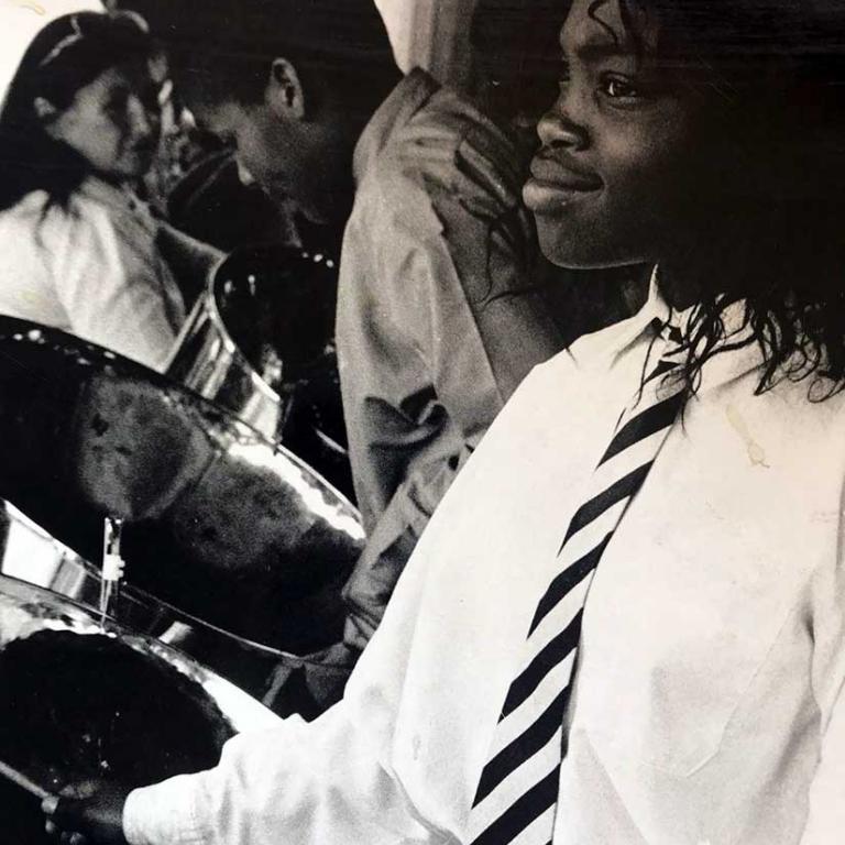 Photo depicts four young people playing steel pans. In the foreground a girl wearing a school tie is smiling as she plays. Photo courtesy of RBKC Local Studies and Archive, circa 1980s.