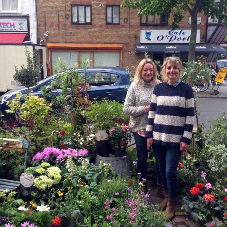 A photo of two ladies smiling at the camera standing beside their flower stall on Golborne Road’s Friday Market. They are surrounded by a wide selection of colourful plants and flowers. In the background is Cafe O’Porto, the beloved Portuguese coffee shop. Photo taken by Darnell Da Costa, 2019.