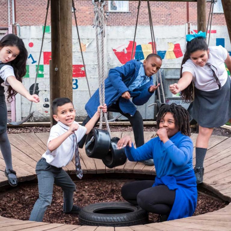 Photo depicts five children dressed in school uniform posing like singers and dancers inside a circular wooden play structure. One of the children holds the ropes of two rubber tyres. In the background is the colourfully painted wall of the younger children’s playground. Photo by Kevin Percival, 2018.