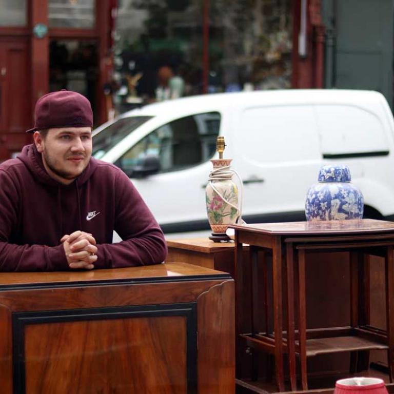 A photo of a young man with a baseball cap leaning on antique mahogany cabinet on the Golborne Road. Beside him are other antiques chairs, tables, a china vase and lamp. In the background is Clarke’s antique shop. Photo taken by Darnell Da Costa, 2019.