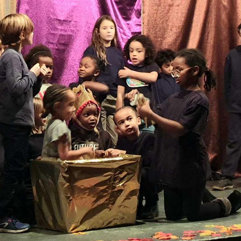 Photo depicts twelve children on the stage of Venture’s main hall. They are from Renegade Theatre’s Drama Club acting in their Christmas play called Cinderella’s Wornington Wish. The child playing Cinderella sits inside a golden box and the other children cluster around her. Shiny purple and brown fabric hangs in the background and silk leaves are scattered at the front of the stage. Photo by Talibah Stevenson, 2019.
