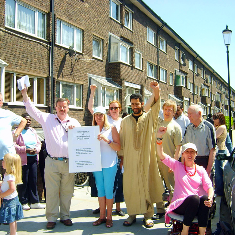 Photo depicts a group of residents and Labour party councillors protesting against the demolition of Wornington Green Estate by declaring independence  from their housing association, Kensington Housing Trust (KHT). They are holding up a constitution of the Republic of Peplar House on a white board. Four residents have raised fists. Photo reproduced from The United Estate of Wornington Green website.