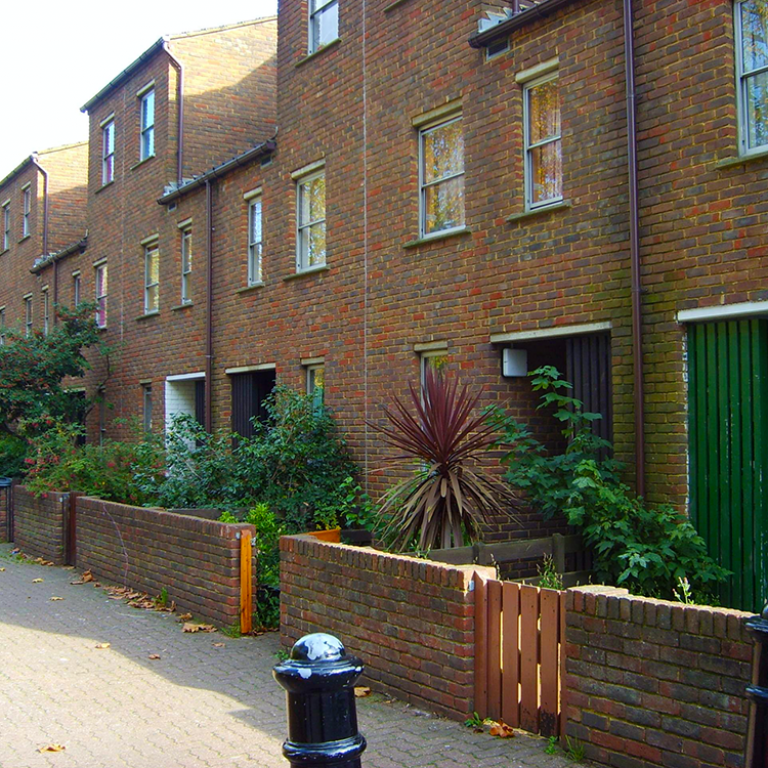 Photo depicts three floor terrace houses on Munro Mews. Each has a small front garden with lush foilage. The street is cobbled with black bollards to restrict car access. Munro Mews was demolished in 2011. Photo reproduced from The United Estate of Wornington Green website.