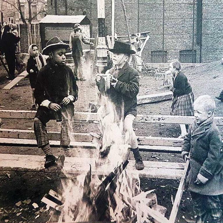 This historical photo is of the original Venture playground. On the left is a boy with a tyre looped on his shoulder standing next to two little boys sitting on the back of a long wooden bench, next to them is a young child in a winter coat and scarf - there is a bonfire infront of them. The background shows other children playing.
