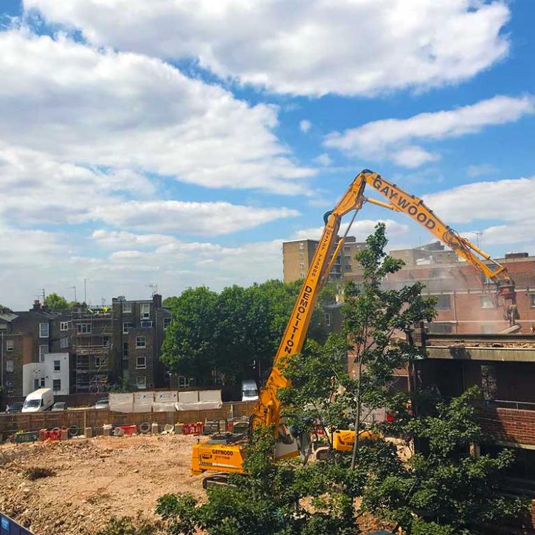 A Gaywood Demolition machine is half way through knocking down the top floor of Murchison House. To the left is the empty building site. The blue sky and white clouds frame the scene. Photo by Sanya Mihaylovic, 2019.