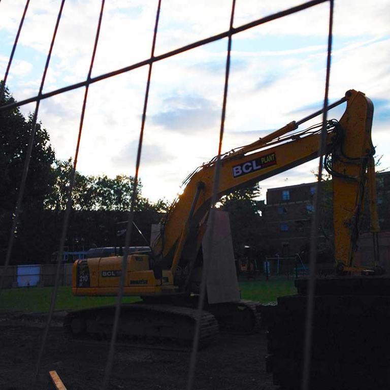 A silhouette photo of an excavator digging on the site of the demolished Lionel House and preparing the ground to create the new Athlone Gardens. The sun sets behind the clouds above. Photo 2014.